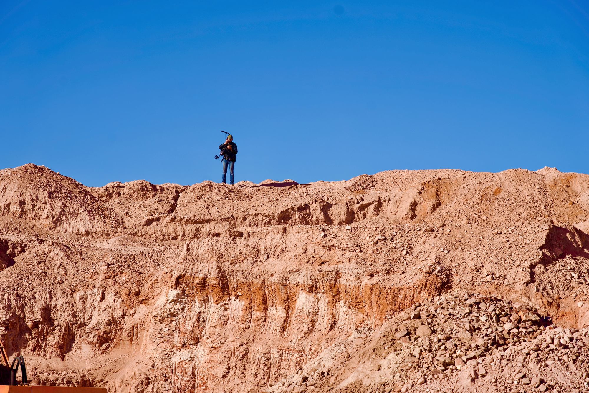 A lone camera operator on a red earth ridge under a blue sky