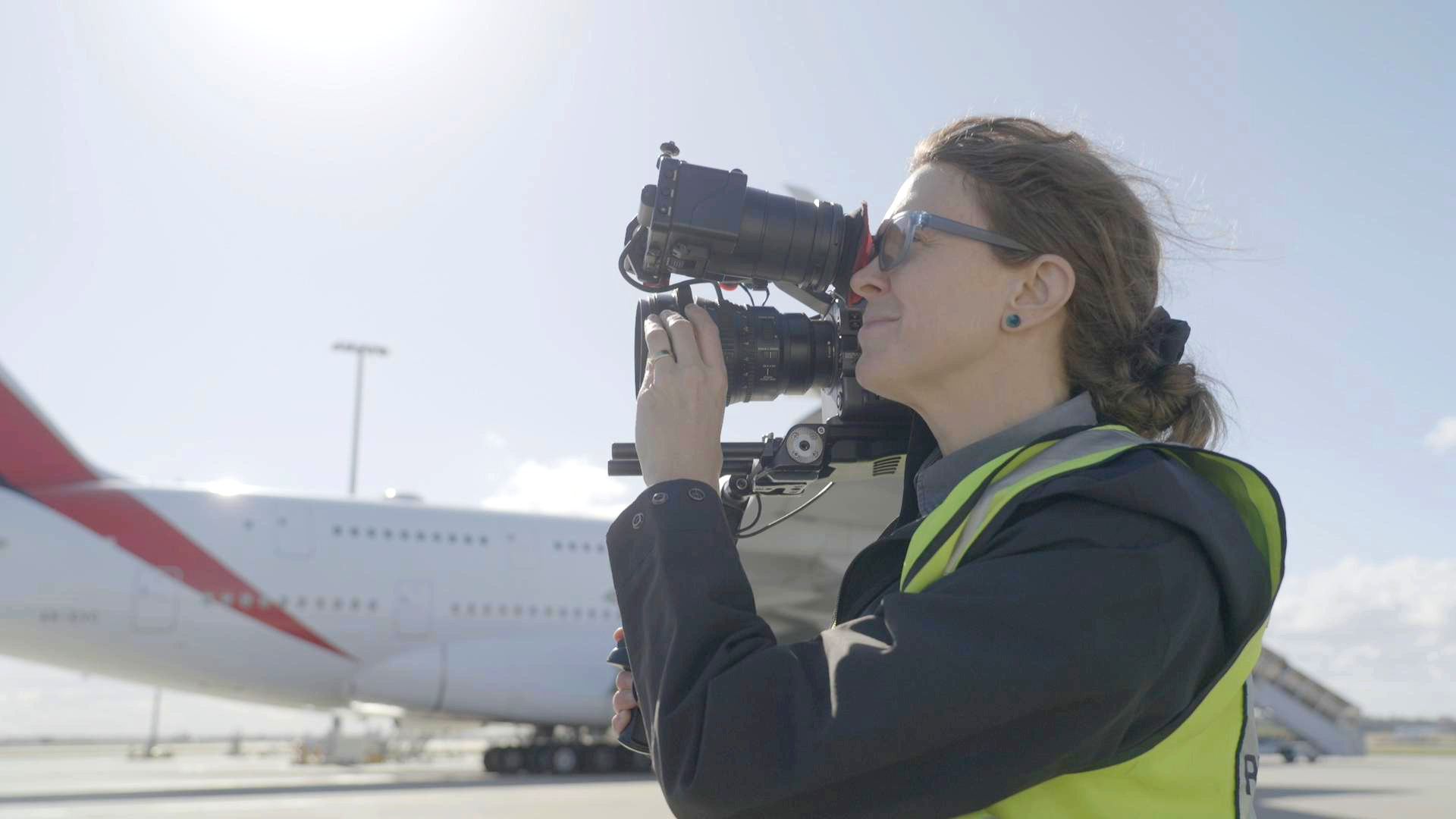 Olivia filming with a shoulder-mounted camera beside a plane